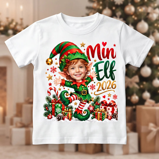 Weißes Kinder-T-Shirt mit personalisiertem Jungen-Elf, grüner Zipfelmütze, Weihnachtsgeschenken und dem Schriftzug „Mini Elf 2026“.