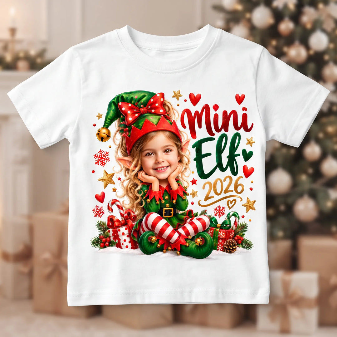 Weißes Kinder-T-Shirt mit personalisiertem Mädchen-Elf, großer weihnachtlicher Schleife und dem Schriftzug „Mini Elf 2026“.